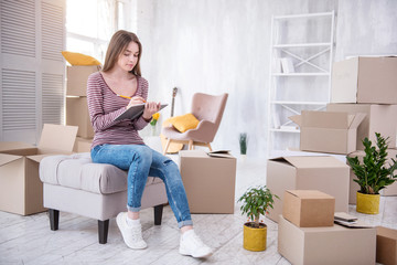 Everything in order. Beautiful young woman sitting on the pouf in her new flat and checking the list of boxes in the notebook