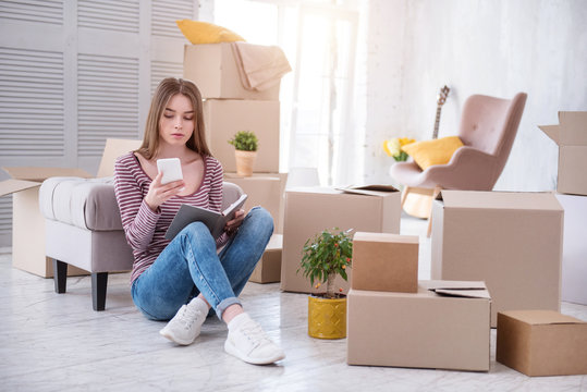 Need assistance. Charming young woman sitting on the floor and checking the number of removers company before moving out of the house