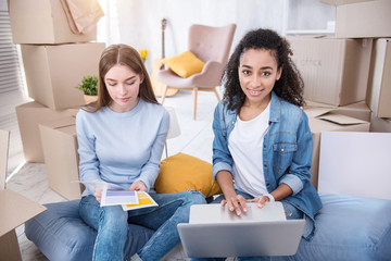 Convenient shopping. Charming female students sitting on the floor among boxes with their belongings and buying paint for new flat online