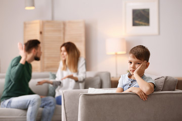 Little unhappy boy sitting in armchair while parents arguing at home