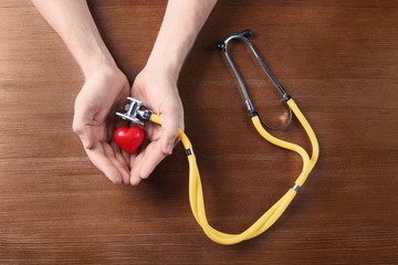 Man holding model of heart and stethoscope on wooden table. Heart attack concept
