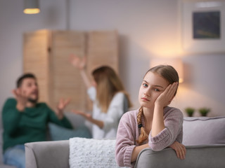 Little unhappy girl sitting in armchair while parents arguing at home