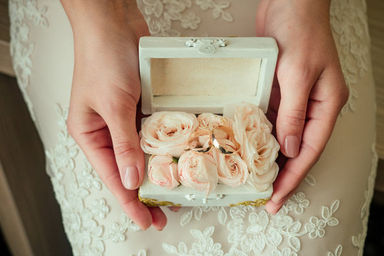 Female hands hold a white box with wedding rings inside. On a background of a white dress of the bride