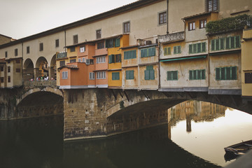 Fototapeta na wymiar Florence, Italy. View of the Golden bridge and old houses on the waterfront of the river Arno. Cloudy sky. Houses are reflected in the water.