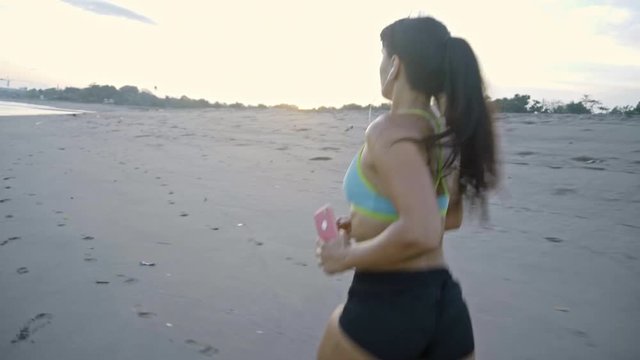 Tracking shot of fit woman listening to music in earphones and running along beach in evening