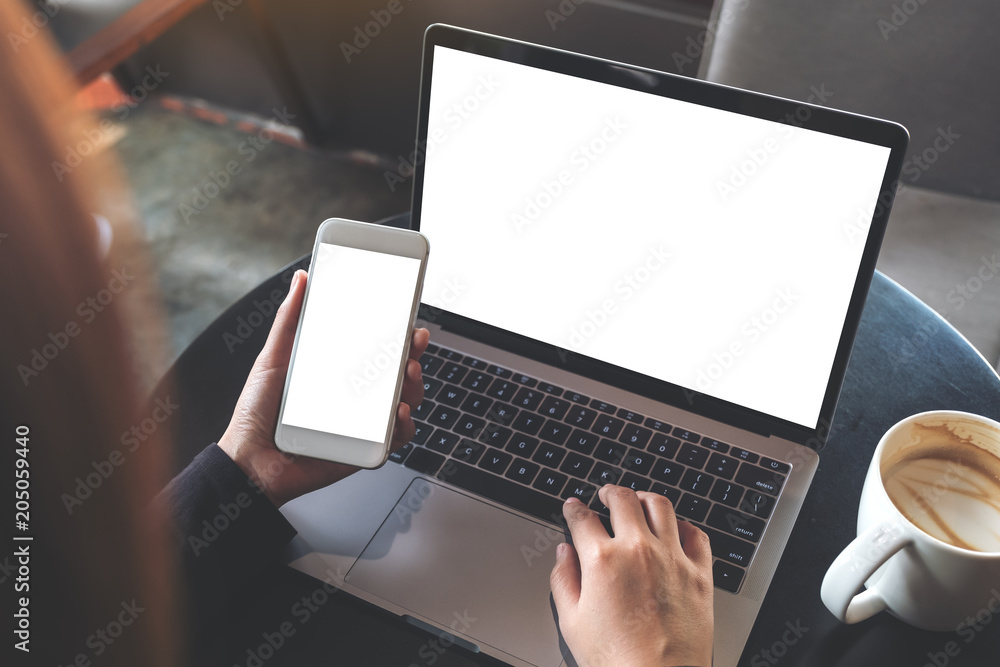 Wall mural top view mockup image of hands holding blank mobile phone while using laptop with blank white deskto
