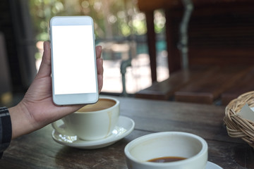 Mockup image of a hand holding and showing a white mobile phone with blank desktop screen with coffee cups on wooden table in cafe