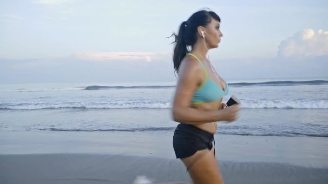 Tracking with tilt up of fit young woman listening to music in earphones and running along beach