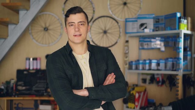 Portrait of bike repair workshop owner standing in his workplace and looking at camera. Bicycle spare parts, reparing equipment and tools are in background.