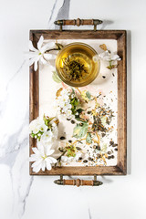 Glass cup of hot green tea on vintage tray with spring flowers white magnolia and cherry blooming branches over white marble texture background. Top view, space.