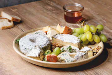 Cheese plate assortment of french cheese served with honey, walnuts, bread and grapes on ceramic plate over wooden table. Close up