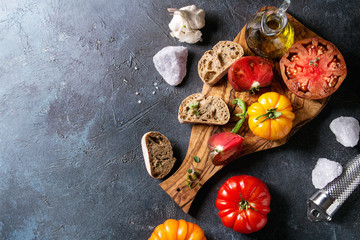 Variety of red and yellow organic tomatoes with olive oil, garlic, salt and bread for salad or bruschetta on wooden cutting board over blue texture background. Top view, space.