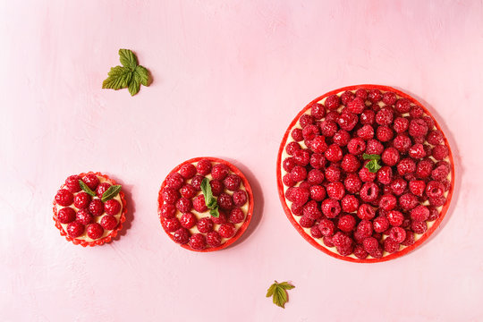 Variety of red raspberry shortbread tarts and tartlets with lemon custard and glazed fresh raspberries over pink pastel background. Top view, space.