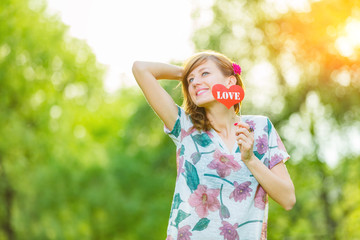 Cute young woman holding a paper heart - shaped sign outdoors.