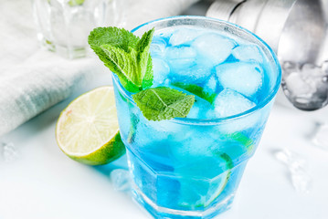 Colorful summer beverage, iced blue lagoon alcohol cocktail drink with lime and mint, white marble...