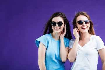 Cute smiling girls dressed in t-shirts and sunglasses listen to the music via earphones before blue background