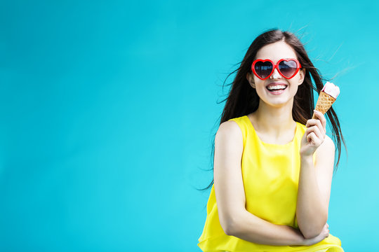 Pretty brunette woman dressed in yellow shirt and sunglasses eating waffle icecream before blue background