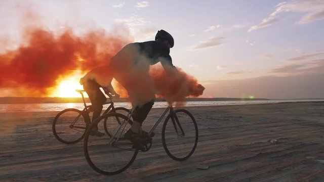 Two young men riding bicycles on the beach with orange smoke during sunset, slow motion