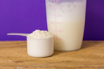 Whey Protein. Front view of scoop with vanilla powder and shaker on rustic wooden table. Solid color: violet.