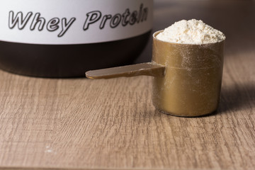Whey Protein. Golden scoop with vanilla flavour powder and black jar of supplement. Wooden table. Close up, texture.