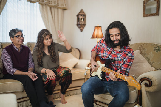 Mid adult man is playing guitar while visiting his parents home. They are sitting on the sofa, dancing and singing along in the background.