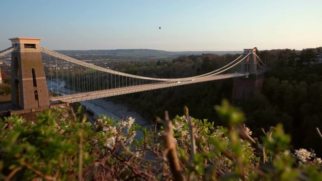 Camera slides over hedgerow with the Clifton Suspension Bridge behind. Shot during a spring sunset with a hot air ballon in the sky