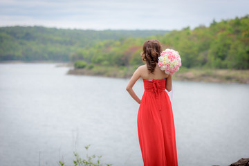 Fototapeta na wymiar Beautiful woman wear red evening dress hold a bouquet of flowers. over mountains and rivers background.