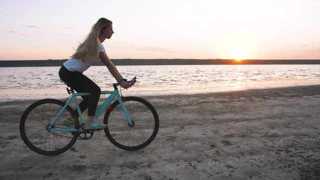 Young attractive woman riding biycle on the beach near the sea during sunrise or sunset, slow motion