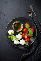 Cherry tomatoes, basil, mozzarella cheese and olive oil in black plate on dark background. Italian food background. Selective focus. Top view.