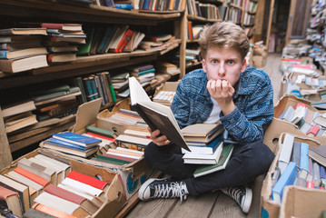 Funny young man sitting on the floor in a public library with a book in his hands and looking at the camera with astonished look. A funny student in a cozy, old library