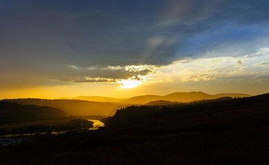 Golden sunset in the mountain valley. dark forest, the river reflects the light of the sun, the blue sky with clouds and rays