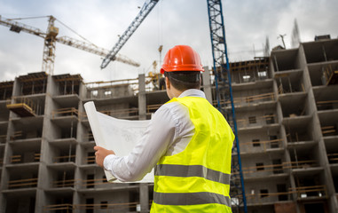 Rear view image of construction engineer looking at blueprints and working cranes on building site