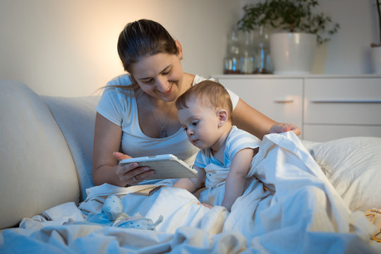 Young Smiling With Baby Boy Watching Cartoons On Digital Tablet Before Going To Sleep