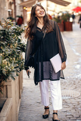 beautiful Hipster Girl in fashionable outfit, smiling and holding magazine in sunny street. happy stylish woman with gorgeous hair and smile, walking in sunlight in city