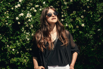 beautiful Stylish Hipster Girl with beautiful hair relaxing at floral bushes in sunny day. Gorgeous Boho Woman with sunglasses in fashionable outfit enjoying day in garden