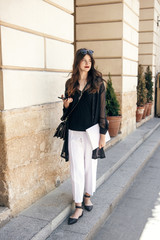 Obraz premium beautiful stylish woman walking in sunny city street, holding phone and magazine. happy hipster girl dressed in fashionable outfit, posing in european city