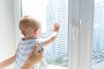 Closeup image of parents hands holding bbay boy standing on windowsill and looking out of window