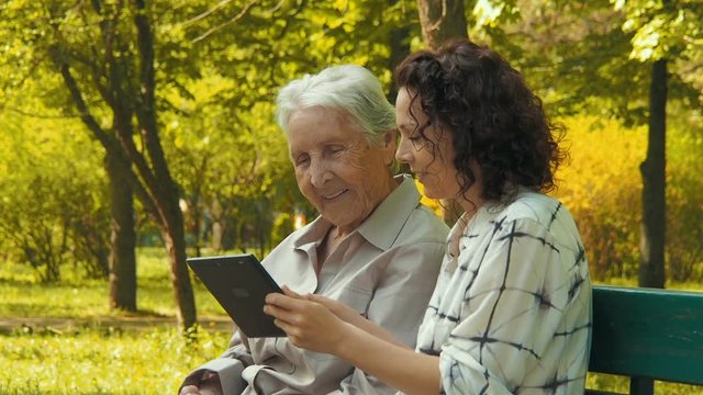 Look at the pictures on the tablet. An elderly woman with a tablet. A woman with an elderly mother with a gadget in the park.