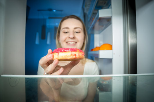 Closeup image of hungry young woman taking high-caloric donut from refrigerator at night. Concept of unhealthy nutrition