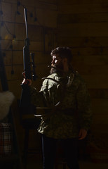 Fototapeta na wymiar Hunter concept. Man with beard wears camouflage clothing, dark background. Macho on strict face at gamekeepers house ready for hunting. Hunter brutal with gun and horns of deer, lighted in darkness.