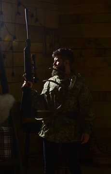 Hunter Concept. Man With Beard Wears Camouflage Clothing, Dark Background. Macho On Strict Face At Gamekeepers House Ready For Hunting. Hunter Brutal With Gun And Horns Of Deer, Lighted In Darkness.