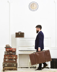 Butler and service concept. Macho attractive, elegant carries vintage suitcases, side view. Man with beard and mustache wearing classic suit delivers luggage, luxury white interior background.