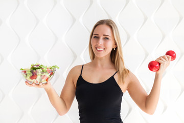 Young fitness model woman athlete in sportswear  with red dumbbells and green salad in glass dish. Healthy life bodybuilding lifestyle concept image, copy space. White empty background for text.