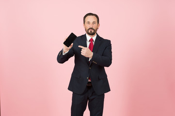 Portrait of young man in black suit pointing finger at blank screen mobile phone isolated over pastel pink background. Look at this smart-phone! Bearded officer pointing by finger to black phone