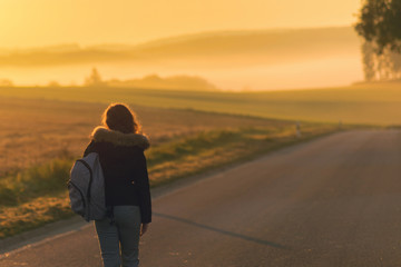 A girl in a jacket with a backpack and suitcase on the road against the background of the autumn field and sunset