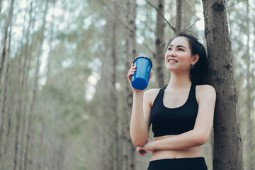 Beautiful asian woman drinking protein shake in the spruce forest nature for relaxing after runner exercise cardio workout or loss of sweat recovery refreshment active healthy lifestyle.