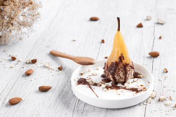 Pear in chocolate and almonds on a white plate, on a wooden white background.