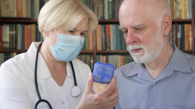 Female professional doctor in medical mask with stethoscope at work. Senior woman physician talking to sick senior male patient at home consulting about new drug. Showing case of pills capsules