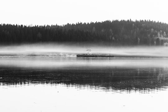 A black and white photograph of how a cargo ship sails in a fog along a river near a coniferous forest