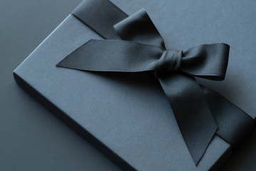 Black gift box on a dark contrasted background, decorated with a textured bow and feathers,...